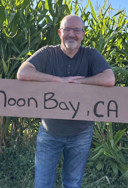 joe is dressed in blue jeans and blue henley top, standing in a cornfield with a sign that reads “half moon bay, CA”