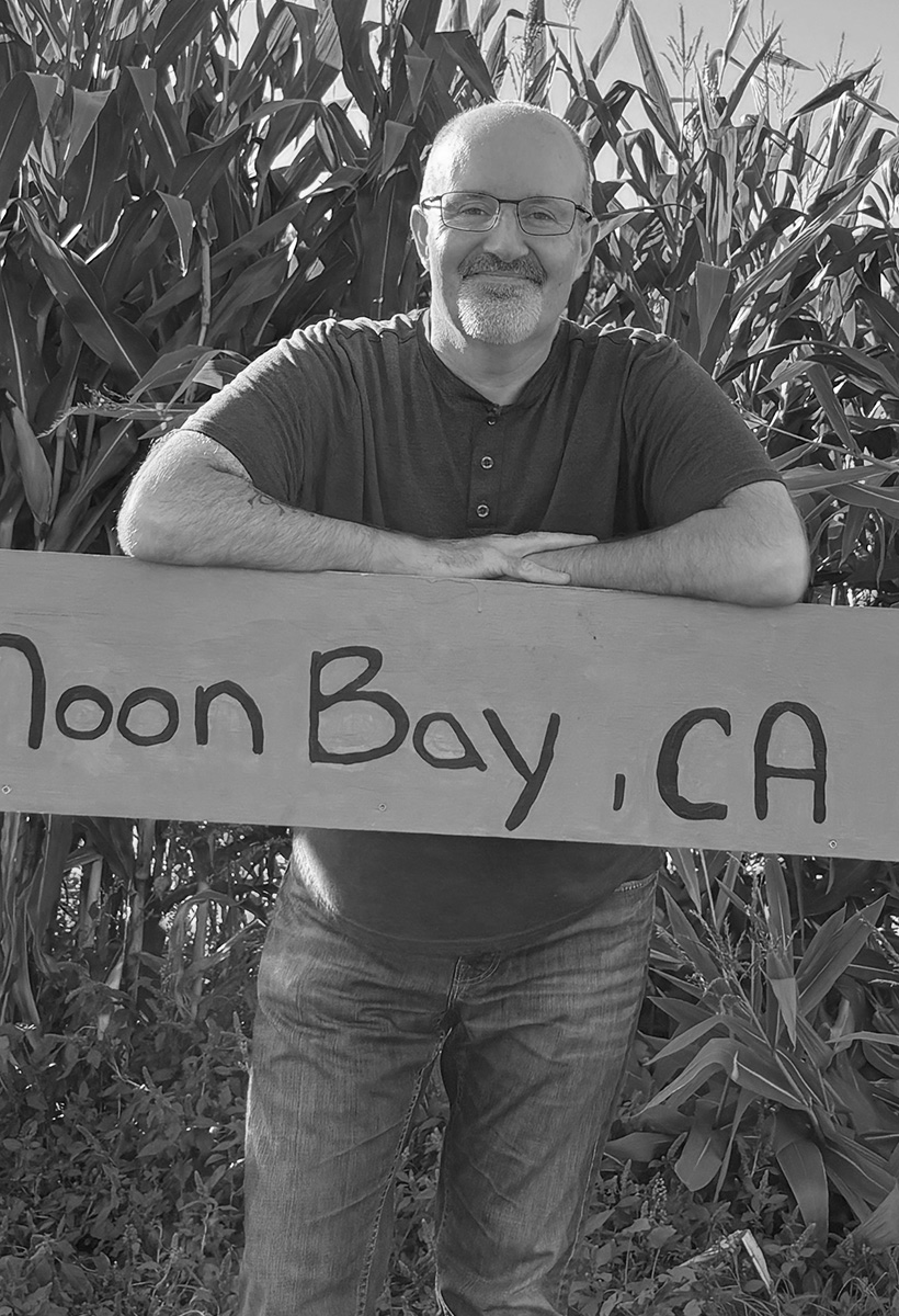 joe antrim is standing in a corn field behind a sign that reads half moon bay, ca. he is balding, wears glasses, and is in a henley shirt and jeans