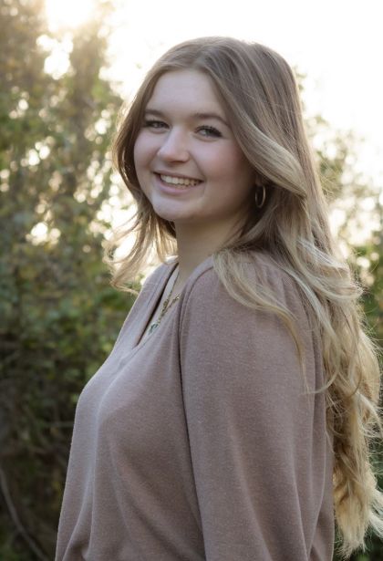 gace is sitting at anoutdoo restaurant under a blue umbrella. she has long brown hair and brown eyes and is weaing a floral sundress.